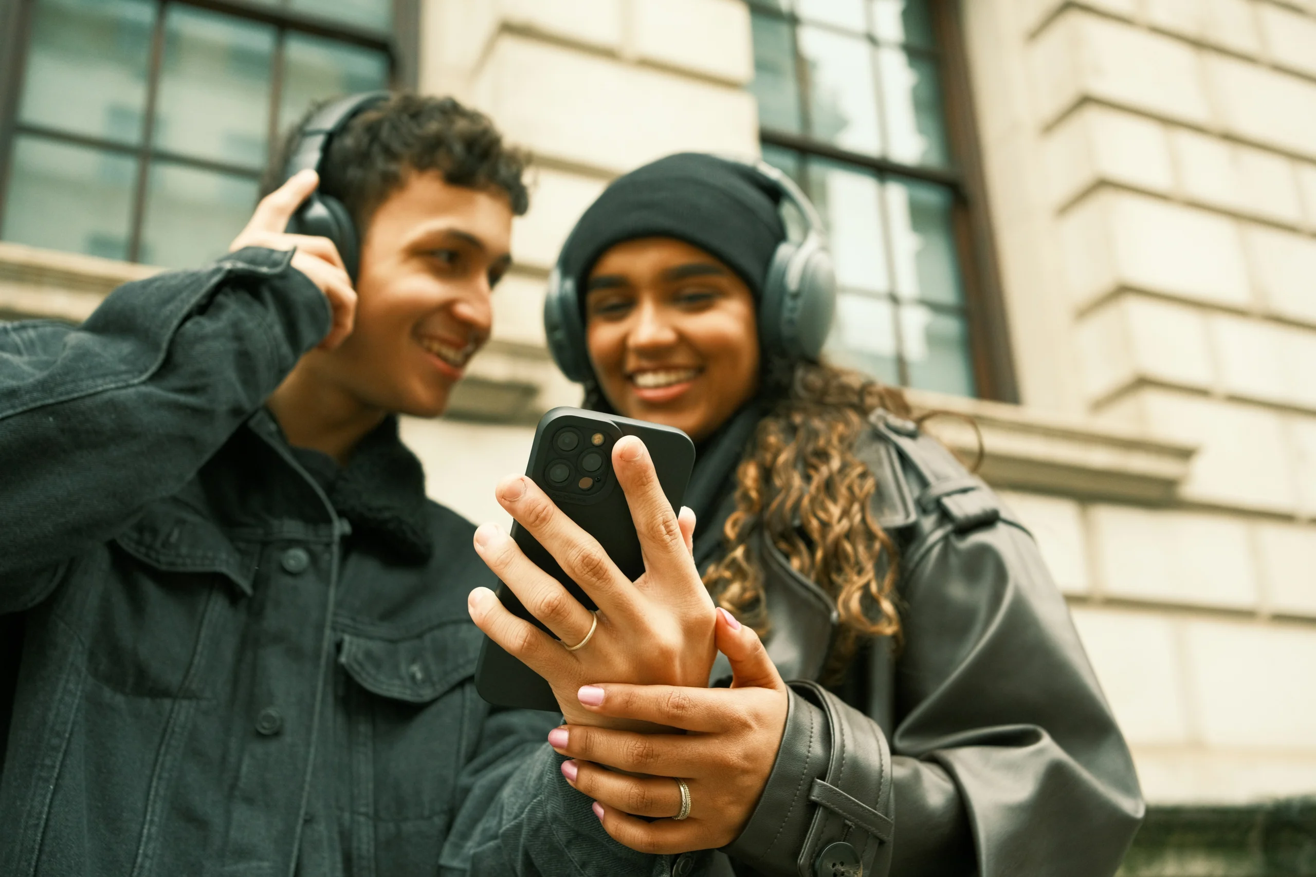 Zwei junge Menschen mit Kopfhörern schauen gemeinsam auf ein Smartphone und lächeln.